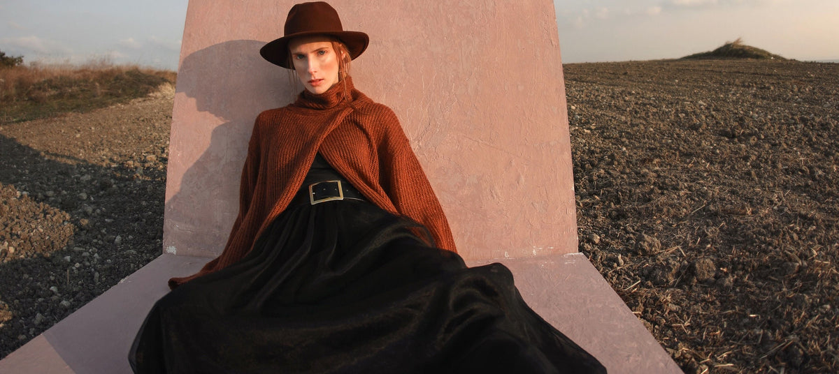 A model wearing a brown wide-brim hat, a rust-brown cape, a long black skirt, and black ankle boots sits on a large concrete sculpture in an open outdoor landscape at sunset.